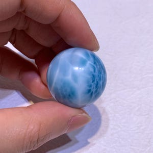 Puede incluir: Una piedra preciosa de larimar pulida y esférica que presenta una superficie lisa con un patrón jaspeado en tonos azul cielo, turquesa y blanco. La piedra muestra inclusiones naturales similares a nubes y vetas azul oscuro.