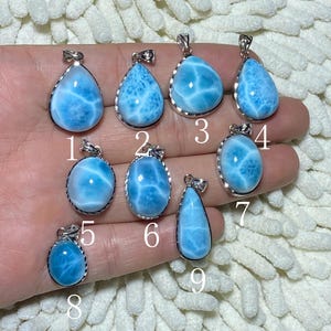 May include: A collection of nine blue larimar gemstone pendants with silver-colored settings. Each pendant has a unique shape, showcasing the stone's natural patterns. The pendants are numbered 1 through 9.