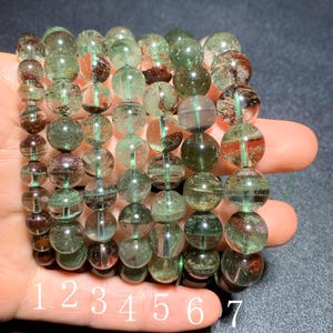 Puede incluir: Varios brazaletes de cuentas de cristal verdes y marrones. Las cuentas son redondas y tienen inclusiones. Los brazaletes están ensartados en un cordón verde. Los números del 1 al 7 están escritos en la mano que sostiene los brazaletes.