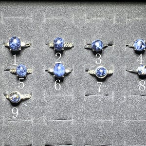 May include: Nine silver rings with blue gemstones are displayed in a black tray. Each ring features a different design, with the gemstones varying in shape and size. The rings are numbered 1 through 9.