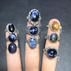May include: A collection of gemstone rings, each featuring a unique oval or round stone in shades of blue, gold, and black. The rings have ornate silver settings, showcasing the variety of designs and stone colors. The rings are displayed on fingers.