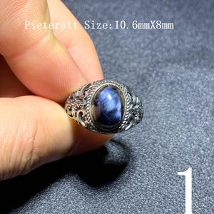 Puede incluir: Anillo de plata con una piedra preciosa ovalada azul. El anillo tiene un diseño ornamental de filigrana alrededor de la piedra. El texto "Pietersit Size: 10.6mmX8mm" es visible en la imagen.