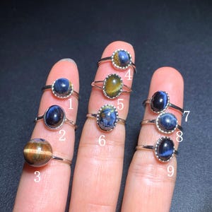 Puede incluir: Una colección de nueve anillos de plata, cada uno con una piedra preciosa diferente. Las piedras preciosas varían en color, incluyendo azul, marrón y amarillo. Los anillos se muestran en dedos sobre un fondo oscuro.