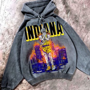 Könnte beinhalten: Grauer Kapuzenpullover mit dem Wort "INDIANA" in gelben und schwarzen Blockbuchstaben. Die Grafik zeigt einen Basketballspieler in Flammen vor einer Skyline. Das Wort "INDIANA" wird am unteren Rand der Grafik wiederholt.