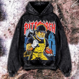 May include: Black hooded sweatshirt with a distressed graphic featuring a construction worker holding a hammer and football, with the word "PITTSBURGH" in flames. The worker wears a hard hat and yellow overalls. The background shows a city skyline.