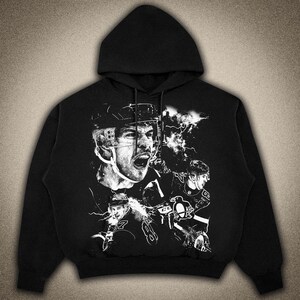 Pode incluir: Hoodie preto com um estampado gr&aacute;fico branco de jogadores de h&oacute;quei vestindo capacetes e uniformes. O gr&aacute;fico inclui o texto "C.B." e um logotipo de pinguim.