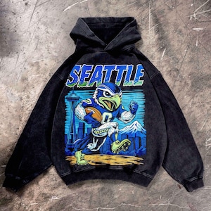 Op de afbeelding: Zwarte hoodie met een afbeelding van een blauw en groen adelaarsmascotte die rent met een voetbal. Het woord "SEATTLE" staat boven de afbeelding. De Space Needle en een bergketen staan op de achtergrond.