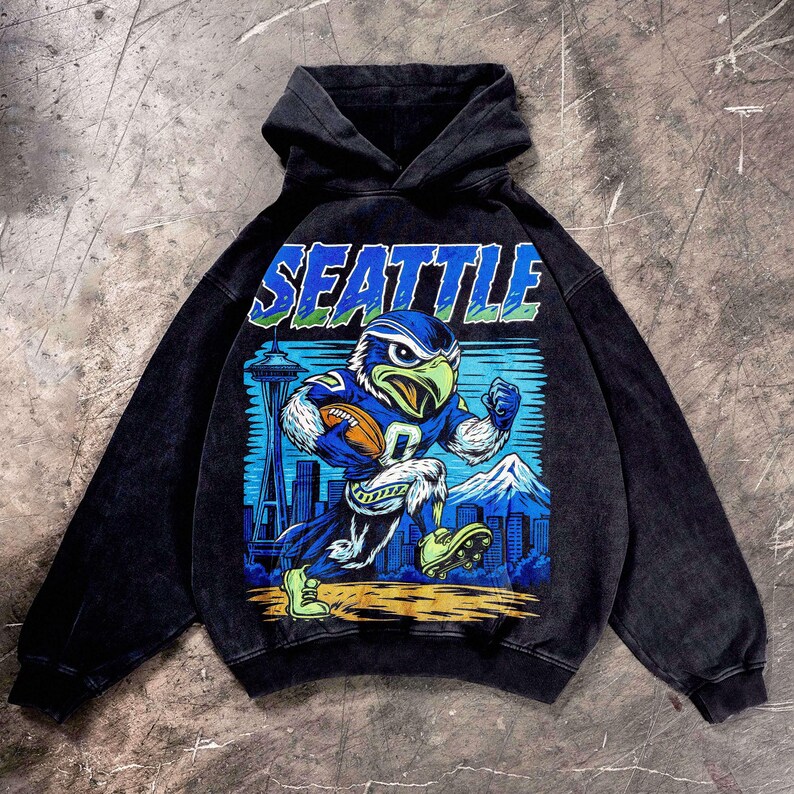 Op de afbeelding: Zwarte hoodie met een afbeelding van een blauw en groen adelaarsmascotte die rent met een voetbal. Het woord "SEATTLE" staat boven de afbeelding. De Space Needle en een bergketen staan op de achtergrond.