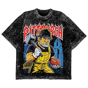 May include: Black acid wash t-shirt featuring a graphic of a construction worker holding a hammer and football, with the word "PITTSBURGH" in fiery letters. The worker wears a hard hat, yellow overalls, and white gloves. A cityscape is in the background.