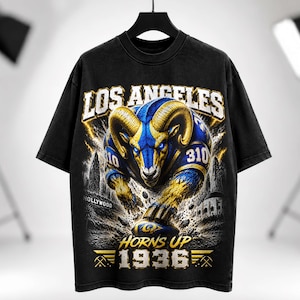 Puede incluir: Una camiseta negra de manga corta con un gráfico de un carnero azul y dorado con grandes cuernos. El texto dice LOS ANGELES, 310, HOLLYWOOD, HORNS UP y 1936. El diseño incluye efectos de rayos y un horizonte urbano de fondo.