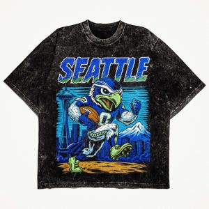 Op de afbeelding: Zwart t-shirt met een afbeelding van een blauwe en groene adelaarsmascotte met een voetbal, met het woord "SEATTLE" in blauw. Het ontwerp bevat een stadsgezicht, bergen en de Space Needle.