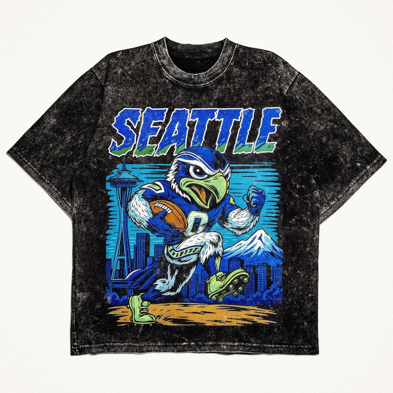 Op de afbeelding: Zwart t-shirt met een afbeelding van een blauwe en groene adelaarsmascotte met een voetbal, met het woord "SEATTLE" in blauw. Het ontwerp bevat een stadsgezicht, bergen en de Space Needle.