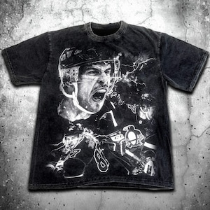 Pode incluir: Camiseta preta com uma estampa gr&aacute;fica branca de um jogador de hockey usando capacete e camisa. O gr&aacute;fico tamb&eacute;m inclui um logotipo de pinguim e raios.