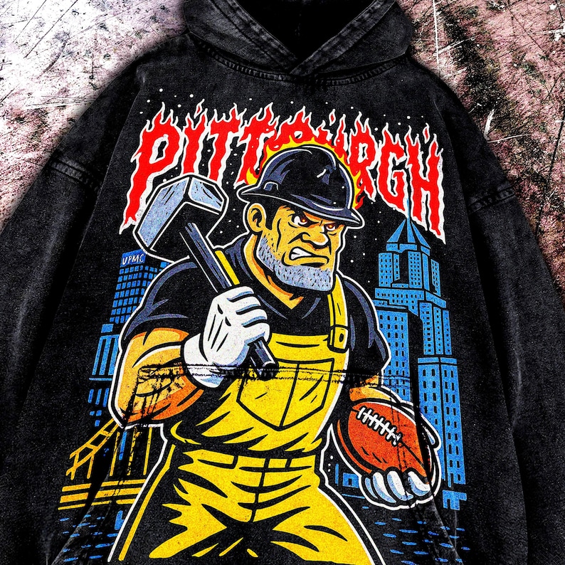 May include: Black hoodie featuring a graphic of a construction worker holding a hammer and football, with the word "Pittsburgh" in red flames. The worker is wearing a hard hat and yellow overalls. The background includes city buildings.