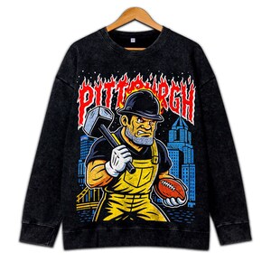 May include: Black sweatshirt with a graphic of a construction worker holding a hammer and football, with the word "PITTSBURGH" in red flames. The worker wears a hard hat, yellow overalls, and white gloves. A city skyline is in the background.