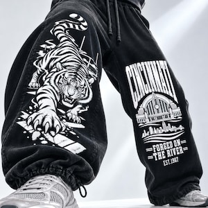 May include: Black denim sweatpants with a drawstring waist and elastic cuffs. The left leg features a large white graphic of a tiger. The right leg displays a white graphic of the Cincinnati skyline with the text "CINCINNATI FORGED ON THE RIVER EST.1967".