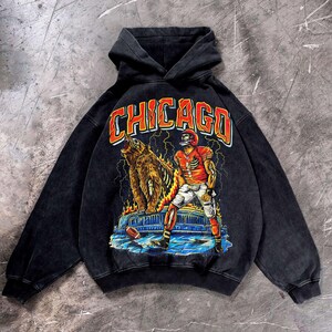 May include: Black hooded sweatshirt with a distressed graphic featuring a bear, football player, and the word "CHICAGO" in orange and white. The design includes flames and lightning bolts, creating a vintage aesthetic.