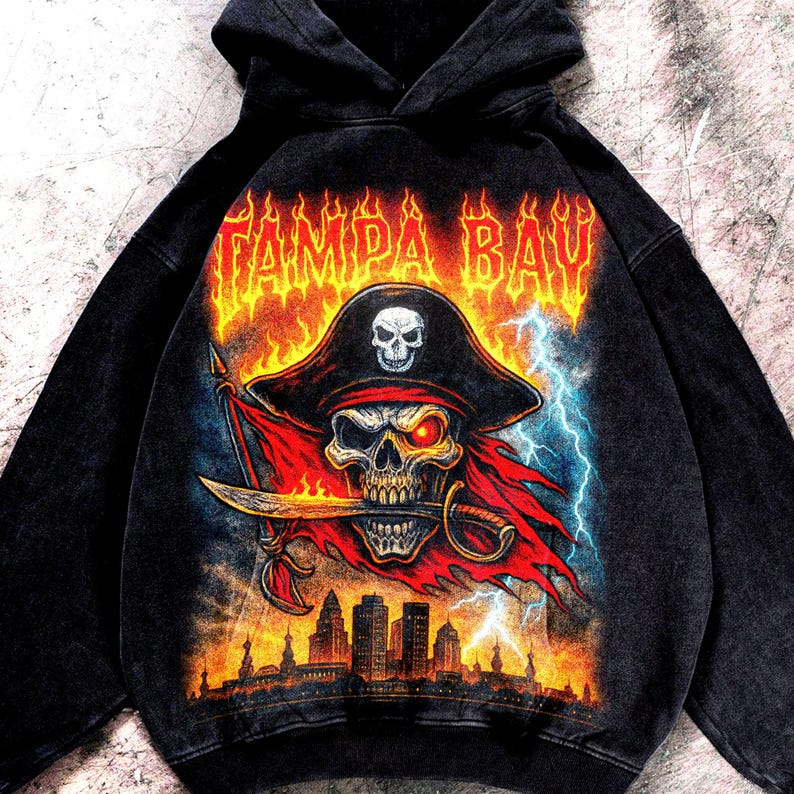 May include: Black hooded sweatshirt with a pirate skull design. The skull has a red eye, a sword in its mouth, and wears a hat with a skull. The words "TAMPA BAY" are in flames above the skull, with a city skyline below.