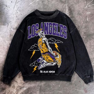May include: Black crewneck sweatshirt featuring a skeleton wearing a basketball uniform with the number 8. The graphic includes the words "LOS ANGELES" and "THE BLACK MAMBA" in purple and white.