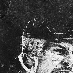 May include: A black and white acid-wash t-shirt featuring a high-contrast graphic print of a hockey player wearing a helmet with a visor. The design captures the detailed texture of the helmet and the player's facial features in a gritty style.