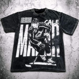 Puede incluir: Camiseta negra con un gráfico blanco de un jugador de hockey que lleva una camiseta de los Colorado Avalanche. El texto "CALE MAKAR" está impreso en la camiseta.