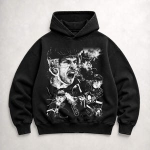 May include: A black hooded sweatshirt featuring a large, detailed white graphic print on the front. The design depicts multiple hockey players in action, including a prominent close-up of a player shouting, alongside a Pittsburgh Penguins logo.