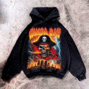 May include: Black hooded sweatshirt with a pirate skull graphic. The design includes a pirate hat, a sword, and the text "TAMPA BAY" in orange flames. The graphic also features a red flag, lightning bolts, and a city skyline.
