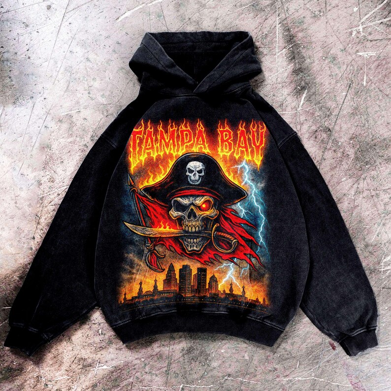 May include: Black hooded sweatshirt with a pirate skull graphic. The design includes a pirate hat, a sword, and the text "TAMPA BAY" in orange flames. The graphic also features a red flag, lightning bolts, and a city skyline.