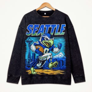 Op de afbeelding: Donkergrijze sweatshirt met een cartoon havik in een voetbaluniform, rennend met een voetbal. Het woord "SEATTLE" staat in blauw en groen boven de havik gedrukt. De Space Needle en een bergketen staan op de achtergrond.