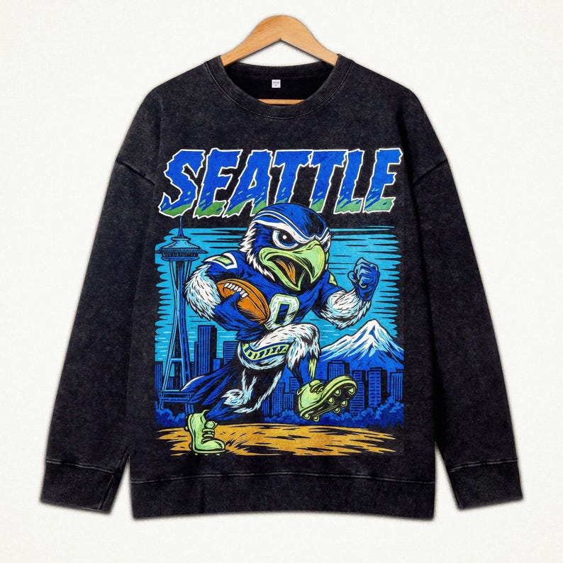 Op de afbeelding: Donkergrijze sweatshirt met een cartoon havik in een voetbaluniform, rennend met een voetbal. Het woord "SEATTLE" staat in blauw en groen boven de havik gedrukt. De Space Needle en een bergketen staan op de achtergrond.