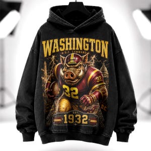 Pode incluir: Um moletom com capuz preto apresentando um gráfico de um javali vestindo um uniforme de futebol americano com o número 32. O texto diz WASHINGTON, D.C. COMMAND e EST. 1932. O fundo inclui relâmpagos e silhuetas do Monumento a Washington e do Capitólio.