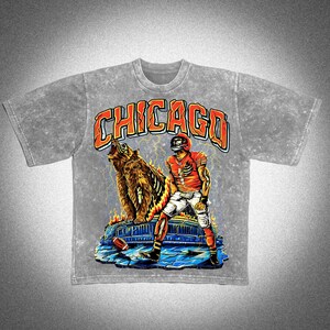 May include: A grey t-shirt with a graphic design featuring the word "CHICAGO" in orange. The design includes a bear, a football player in a red uniform, and lightning bolts. The t-shirt has short sleeves and a crew neck.