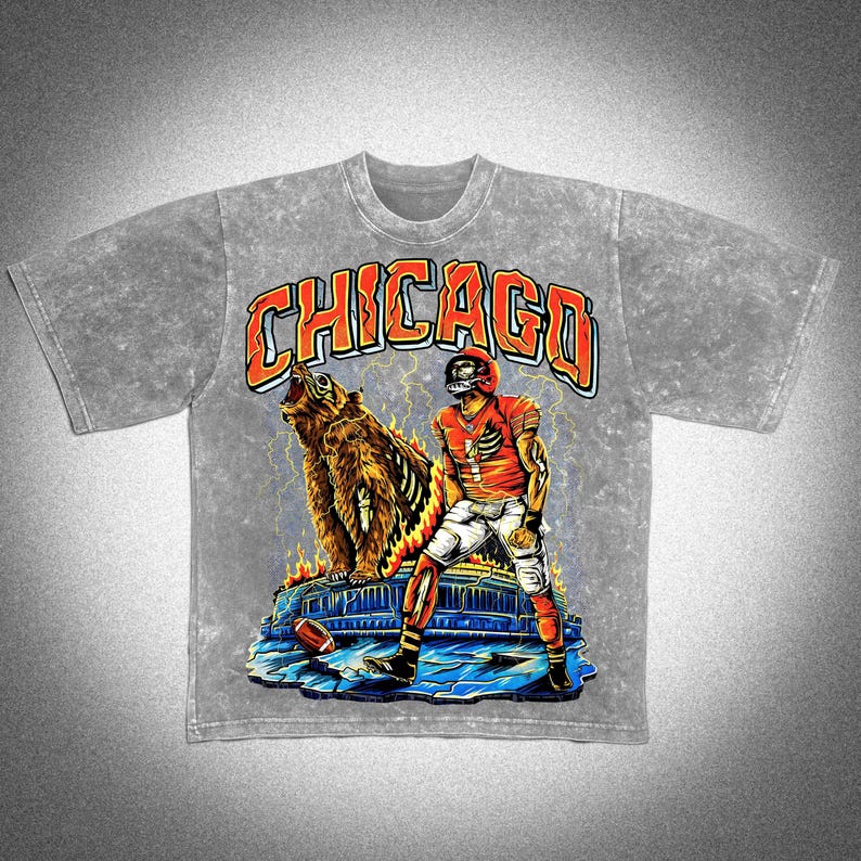 May include: A grey t-shirt with a graphic design featuring the word "CHICAGO" in orange. The design includes a bear, a football player in a red uniform, and lightning bolts. The t-shirt has short sleeves and a crew neck.