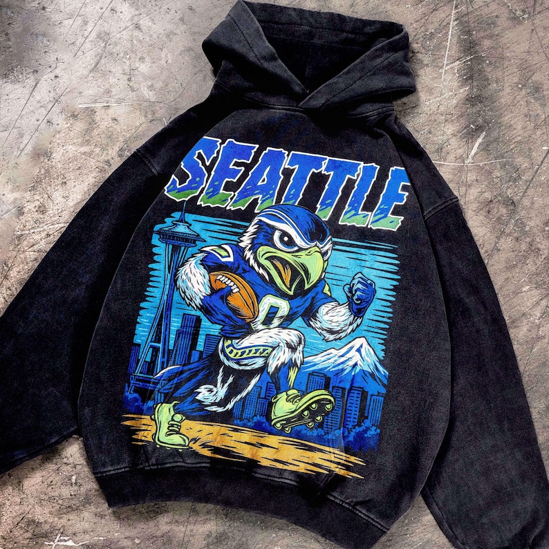 Op de afbeelding: Zwarte hoodie met een afbeelding van een American football spelende adelaar en het woord "SEATTLE". De adelaar draagt een blauw en groen uniform en houdt een voetbal vast. De achtergrond toont een stadsgezicht en bergen.