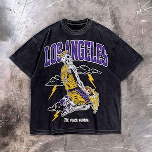 May include: Black t-shirt featuring a skeleton in a basketball uniform with the number 8, a halo, and the words "LOS ANGELES" in purple. The phrase "THE BLACK MAMBA" is printed below the graphic. The shirt has a vintage wash.