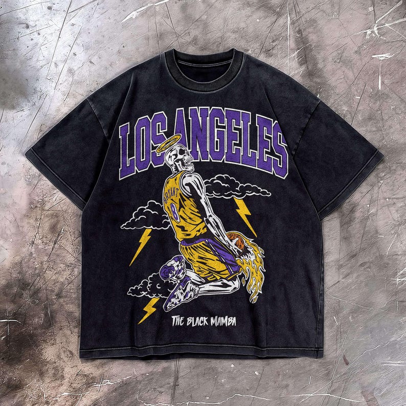 May include: Black t-shirt featuring a skeleton in a basketball uniform with the number 8, a halo, and the words "LOS ANGELES" in purple. The phrase "THE BLACK MAMBA" is printed below the graphic. The shirt has a vintage wash.