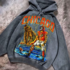 May include: A grey hooded sweatshirt with a graphic design. The design features a football player and a bear, with the word "CHICAGO" in orange and red. The design includes lightning bolts and flames, with a football on the bottom.