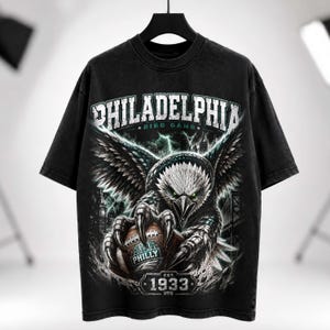 Peut inclure: Un t-shirt noir à manches courtes avec un motif représentant un aigle tenant un ballon de football américain. Le texte indique PHILADELPHIA, BIRD GANG, PHILLY et EST. 1933. Le design inclut des éclairs, une ville et un pont au style vintage.