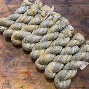 Puede incluir: Varios ovillos de hilo en tonos amarillos, azules y blancos están dispuestos sobre una superficie de madera. El hilo parece estar teñido a mano, con sutiles variaciones de color. Los ovillos están cuidadosamente enrollados, listos para tejer o hacer ganchillo.