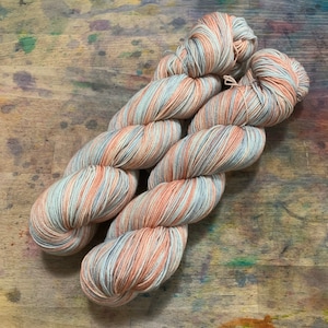 May include: A skein of hand-dyed yarn in shades of peach, gray, and cream. The yarn is twisted into a loose braid, showcasing the color variations. The background is a wooden surface with colorful paint splatters.