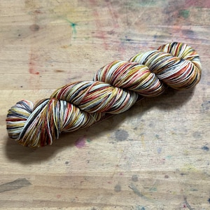 May include: A skein of multi-coloured yarn, featuring red, yellow, white, black, and brown strands. The yarn is twisted, suggesting it's for knitting or crochet. The yarn is displayed on a wooden surface.