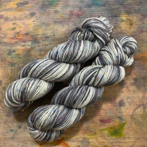 Puede incluir: Dos madejas de hilo jaspeado en tonos grises, blancos y negros. El hilo tiene motas de colores y está dispuesto sobre una superficie de madera con salpicaduras de pintura. El hilo es probablemente para proyectos de tejido o ganchillo.