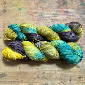 Könnte beinhalten: Ein Strang handgefärbter Wolle in Gelb-, Türkis- und Lilatönen. Das Garn ist zu einem lockeren Zopf gedreht, der die Farbvariationen zeigt. Das Garn besteht wahrscheinlich aus Wolle oder einer ähnlichen Faser, ideal für Strick- oder Häkelprojekte.