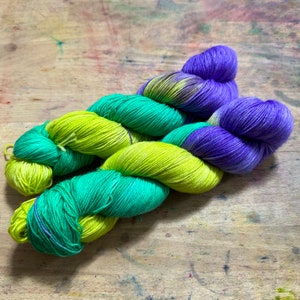 May include: Skein of hand-dyed yarn in vibrant colors. The yarn features sections of bright green, teal, and deep purple. The yarn is arranged on a wooden surface, showcasing the color variations and texture.