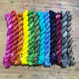 May include: A collection of ten skeins of yarn in a rainbow of colors. The yarn is tightly wound and arranged in a row, transitioning from pink to orange, yellow, green, blue, and purple. The yarn is on a wooden surface.