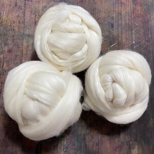 May include: Three white, fluffy fiber balls are arranged on a dark, textured wooden surface. The fiber appears soft and has a slight sheen, suggesting a natural material like wool or silk. The balls are tightly wound, showcasing the texture of the fiber.