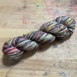 Puede incluir: Un ovillo de hilo jaspeado en una variedad de colores, incluyendo rojo, amarillo, negro y blanco. El hilo está torcido y parece estar hecho de una fibra natural y suave. El hilo está sobre una superficie de madera.