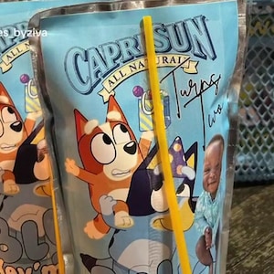 Puede incluir: Bolsas de zumo Capri Sun azules con los personajes de dibujos animados Bluey y Bingo y una pajita amarilla. Las bolsas tienen el texto "CAPRI SUN ALL NATURAL" y "Turns Two". El diseño está sobre un fondo azul claro.