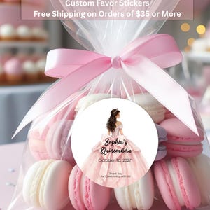 May include: A clear plastic bag filled with pink and white macarons, tied with a pink satin ribbon. A round sticker on the front features an illustration of a person in a pink ballgown with the text: Sophia's Quinceañera, October 10, 2027, Thank You for Celebrating with Us!
