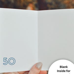 May include: A white greeting card featuring the number 50 printed in a dark blue outline font on the bottom left corner. The interior of the card is blank, providing space for a personal message. The card is held open to display the interior surface.